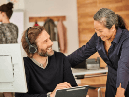 Kollegin und Kollege unterhalten sich am Arbeitsplatz, während der Kollege mit Headset auf seinem Tablet arbeitet