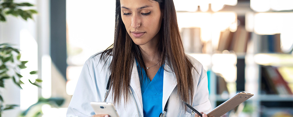 Eine Frau in medizinischer Berufskleidung hält einen Stift und Notizblock in einer Hand, während sie auf ihr Handy in der anderen Hand blickt