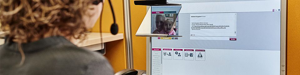 Digitaler Bürgerservice: Eine Frau sitzt mit Headset an einem Büroschreibtisch und schaut auf einen Bildschirm, auf dem ein Videoanruf läuft