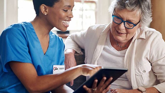 Eine Pflegerin des AWO Schwaben e.V. in blauer Arbeitskleidung nutzt die WLAN-Lösung und erklärt einer älteren Dame Dinge am Tablet