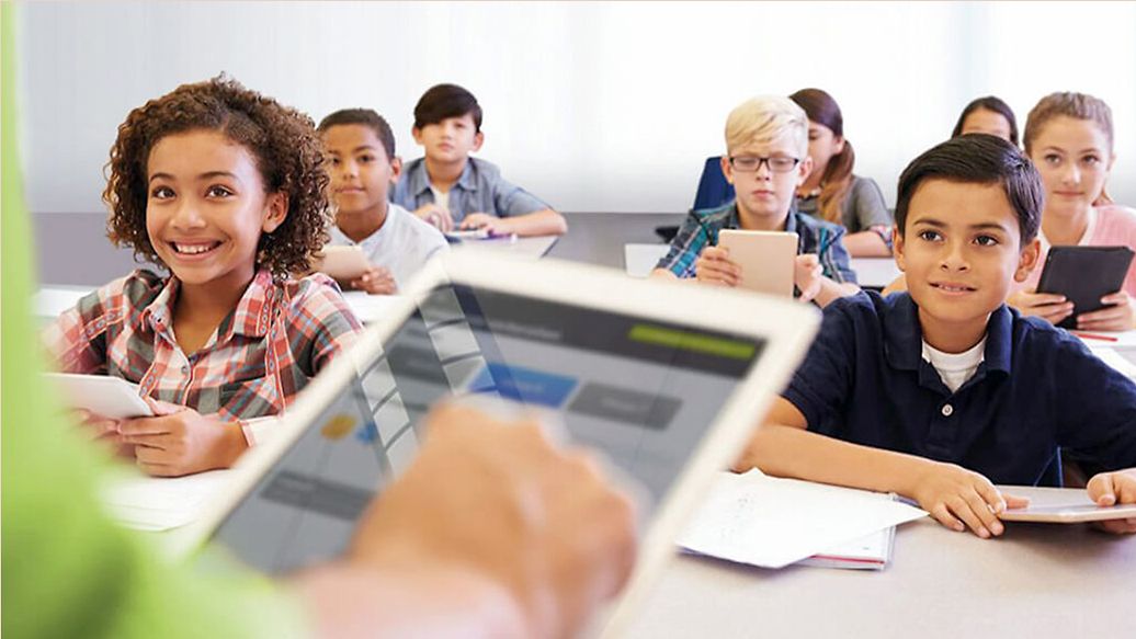 Klassenraum mit lächelnden Schülern und davor ist eine Hand auf einem Tablet zu sehen