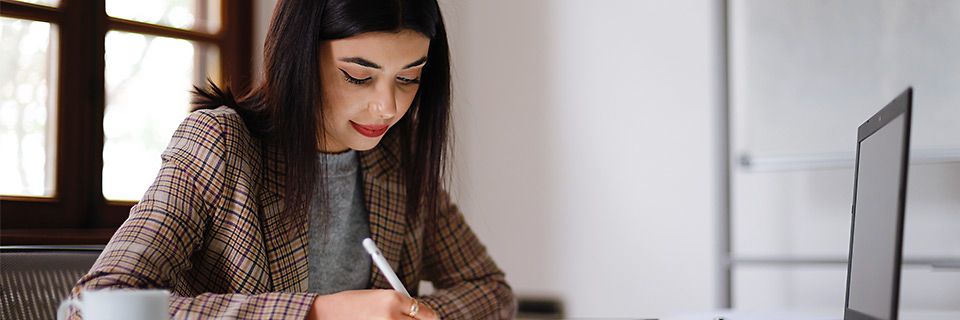 Eine Frau sitzt am Schreibtisch und schreibt mit Pen auf einem Tablet – vor ihr steht ein Laptop