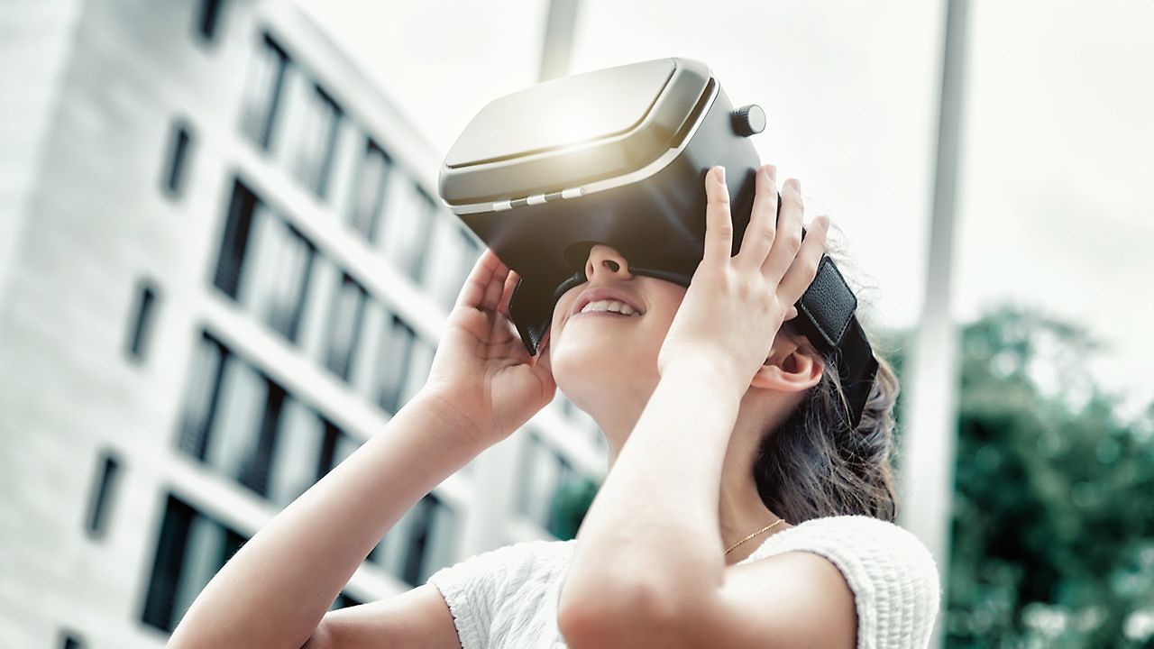 Schülerin benutzt eine VR Brille. Im Hintergrund sieht man schemenhaft ein Schulgebäude.