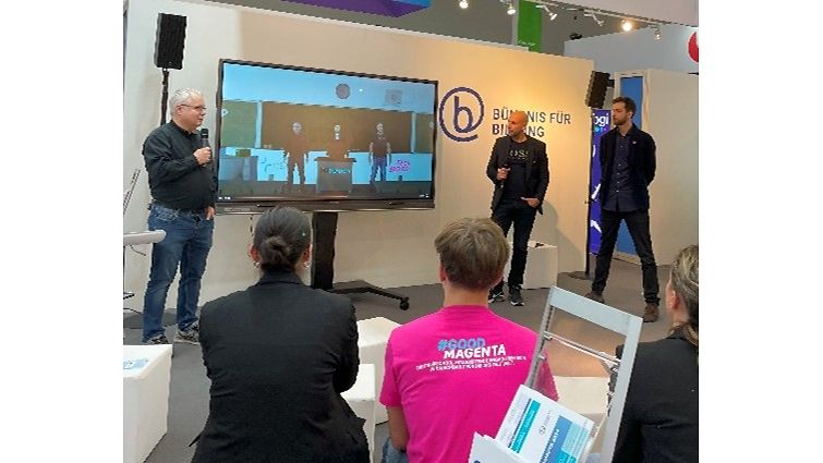 Eine Präsentation an einem Bildungsstand mit Zuschauern, darunter einer in magenta-farbigem T-Shirt