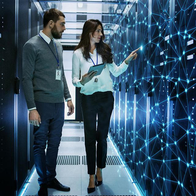 Zwei Personen gehen durch einen Serverraum und betrachten digitale Visualisierungen an der Wand