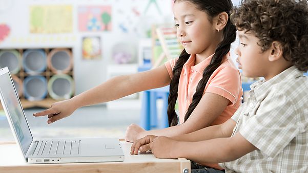 Zwei Kinder arbeiten im Klassenzimmer gemeinsam an einem Laptop, ein Mädchen zeigt auf den Bildschirm.