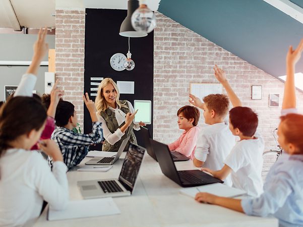 Lehrerin zeigt im Klassenzimmer ein Tablet, während mehrere Kinder mit Laptops sitzen und sich melden