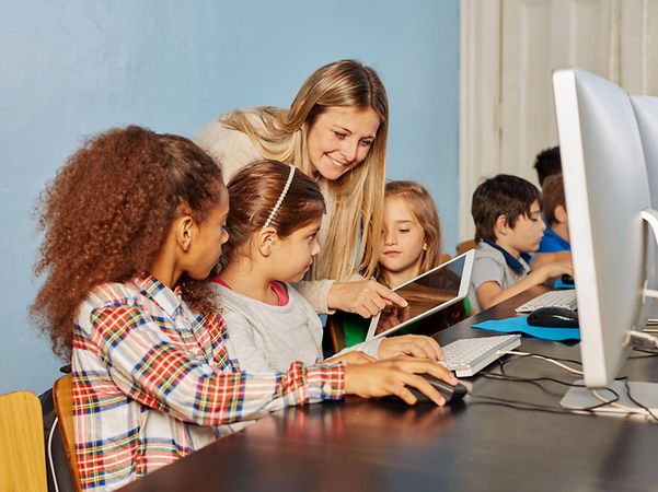 Kinder arbeiten gemeinsam mit Tablets und einem Computer im Unterricht, unterstützt von einer Lehrkraft