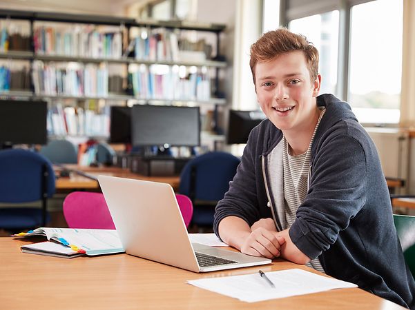 Person sitzt in einem Bibliotheksraum an einem Tisch und arbeitet mit einem Laptop und Notizmaterial