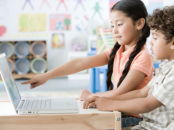 Two children are working together on a laptop in the classroom; a girl is pointing at the screen.