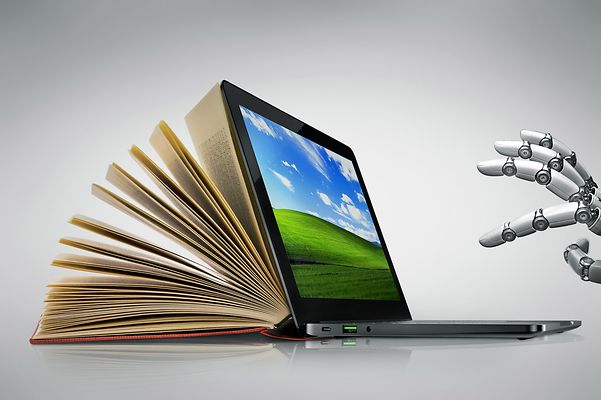 Aufgeschlagenes Buch neben einem Laptop, während eine robotische Hand den Laptop berührt