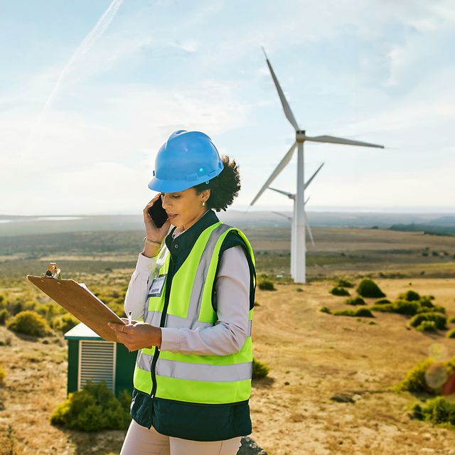 Frau mit Schutzhelm und Warnweste telefoniert mit Notizblock in einer Landschaft mit Windrädern.
