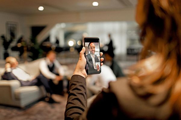 Person hält ein Smartphone mit Videotelefonat, im Hintergrund sitzen mehrere Personen in einem Raum