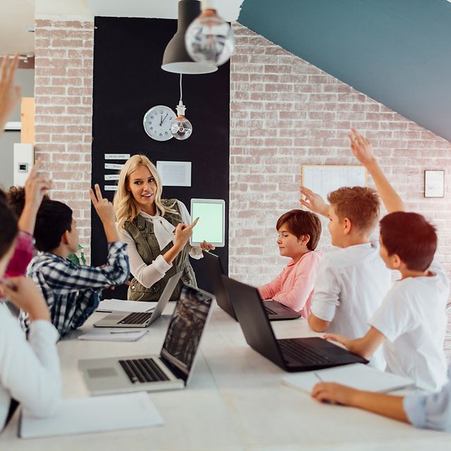 A teacher is demonstrating a tablet in the classroom, whilst several children sit with laptops and raise their hands
