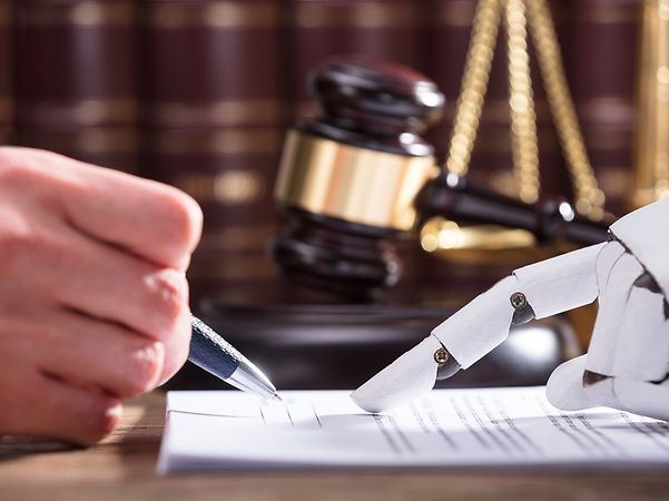 A human hand and a robotic hand sign a document, with a judge’s gavel and law books in the background