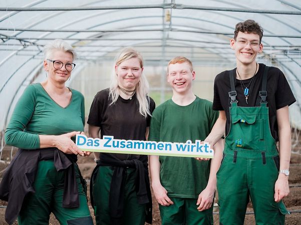 Vier Personen stehen in Arbeitskleidung in einem Gewächshaus und halten ein Schild mit der Aufschrift „Das Zusammen wirkt.“