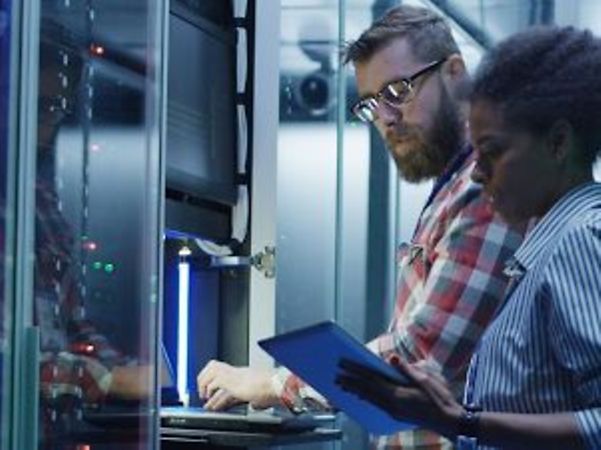 Zwei Personen arbeiten gemeinsam in einem Serverraum an IT-Systemen und prüfen Daten auf einem Tablet.