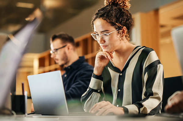 A person is working on a laptop in the office, with another person in the background.
