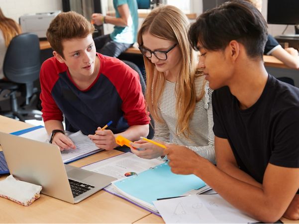 Three young people are working together at a table, using a laptop, documents and pens.