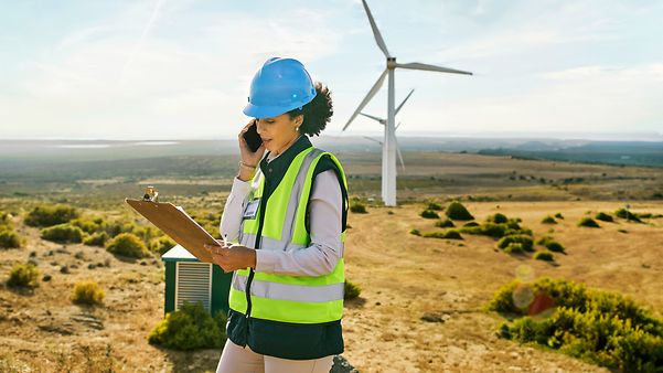 Frau mit Schutzhelm und Warnweste telefoniert mit Notizblock in einer Landschaft mit Windrädern.