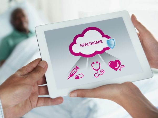 Two people are holding a tablet displaying digital healthcare icons in front of a hospital bed.