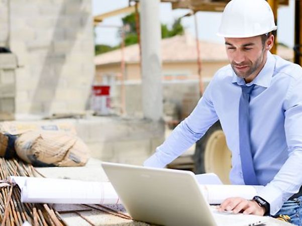 A person wearing a hard hat is working on a laptop next to some building plans on a building site.