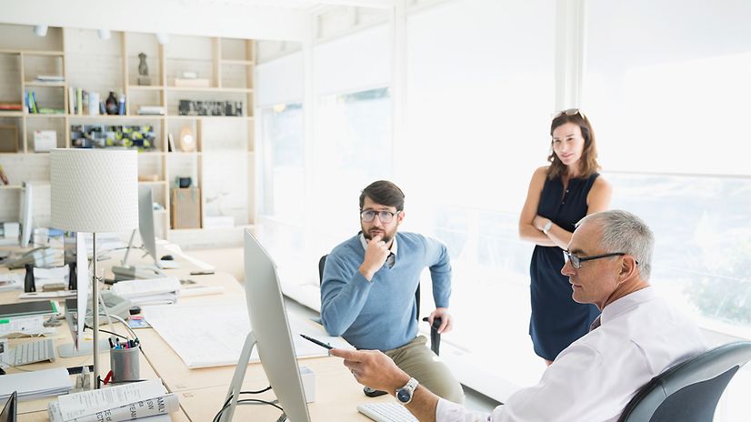 Three people are talking to each other at a computer in a bright office.