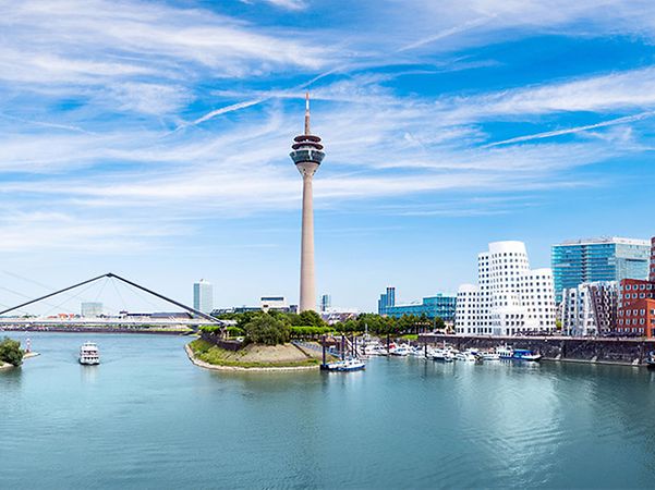 Blick auf den Düsseldorfer MedienHafen mit Rheinturm, moderner Architektur, Yachthafen und Schiff bei blauem Himmel.